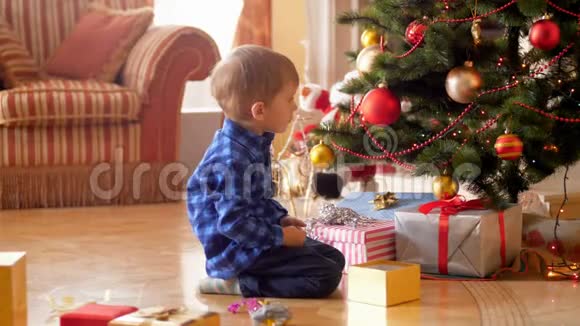4k画面小男孩坐在圣诞树旁的地板上看着闪闪发光的圣诞灯和圣诞饰品视频的预览图