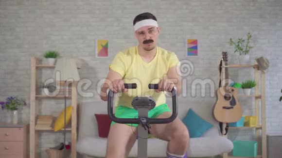 一位80留着胡子在家里骑着一辆静止自行车的精力充沛的男子运动员视频的预览图