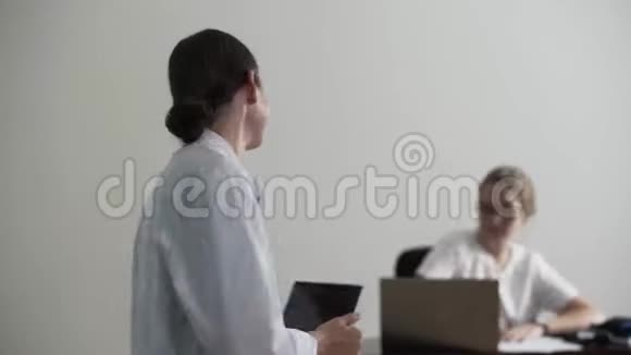 快乐喜悦的年轻职业女医生一边拿着笔记本电脑一边看着镜头在后台坐着一个女孩视频的预览图