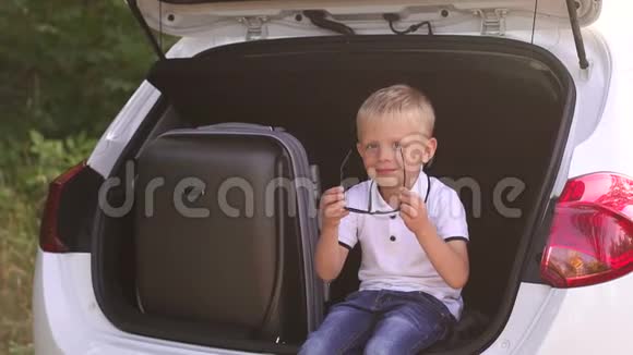 小男孩戴上太阳镜坐在一辆装有手提箱的敞篷车的后备箱里视频的预览图
