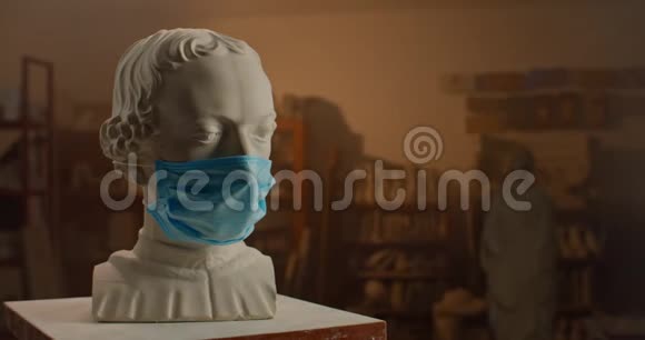 戴着医疗面具的名人的白色石膏雕塑头站在雕塑家工作室的小桌子上被征服视频的预览图