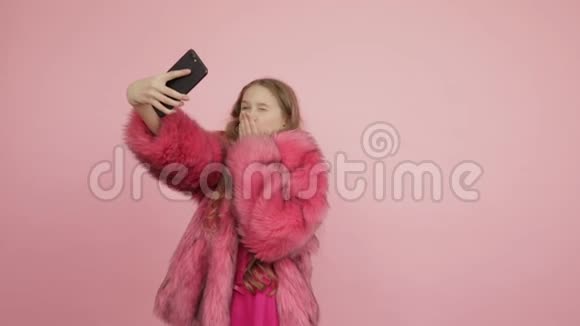 年轻漂亮的女孩穿着粉红色的毛皮外套在粉红色的背景上需要很多自拍并发送空气吻视频的预览图