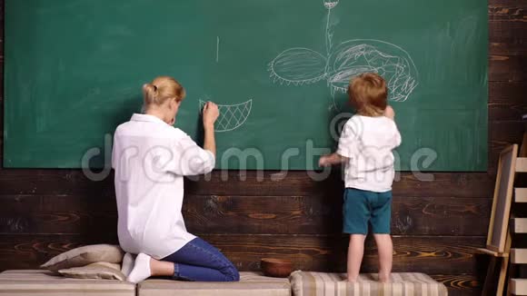 妈妈和她的儿子在学校用粉笔在绿色黑板上画画后视女人和男孩画画回到学校视频的预览图