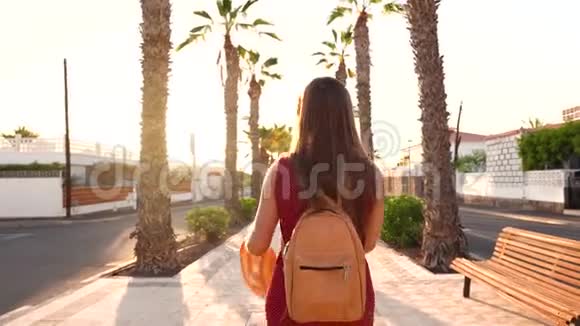 日落时分戴着一顶黄色大帽子的幸福女人沿着棕榈巷散步背面视频的预览图