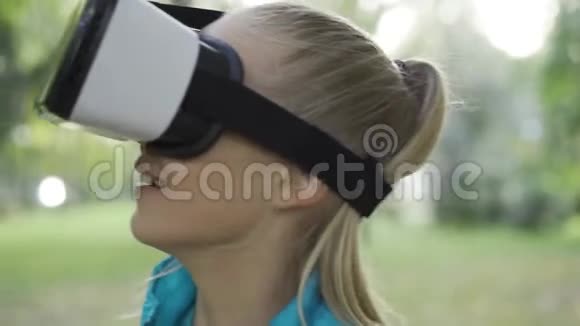 戴着虚拟现实眼镜的金发白人少女在阳光明媚的公园里消磨时光戴虚拟护目镜的积极青少年视频的预览图