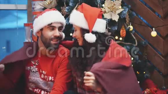 幸福的男人和女人坐在圣诞树附近裹着红色的毯子摸着鼻子视频的预览图