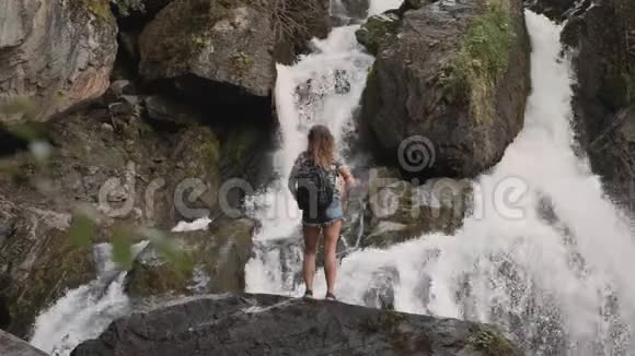 一位背着背包的女游客举起双手站在悬崖边上的一个大瀑布对面快乐视频的预览图