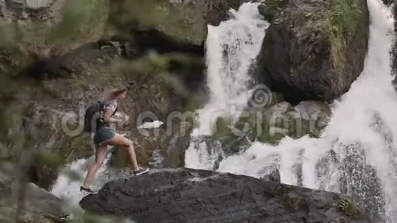 一位女游客爬上一块岩石在一个大瀑布前举起双手快乐的女孩成功了视频的预览图