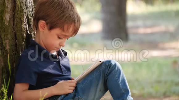 学龄前可爱男孩的肖像孩子坐在公园的一棵树上花时间在平板电脑里玩视频的预览图