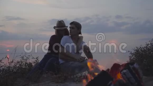 年轻夫妇一边拥抱一边坐在傍晚五颜六色的日落和篝火附近的夏日海滩的背景上视频的预览图