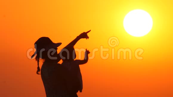母亲戴着帽子带着孩子看着夕阳婴儿在母亲手上的橙色天空背景剪影式视频的预览图