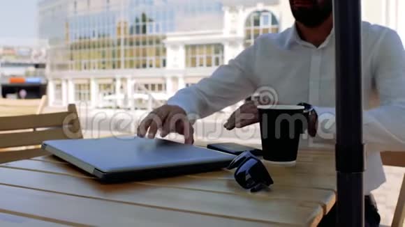 一位留着白衬衫胡子的迷人年轻人坐在街上在咖啡馆里打开一台笔记本电脑喝了一口茶视频的预览图