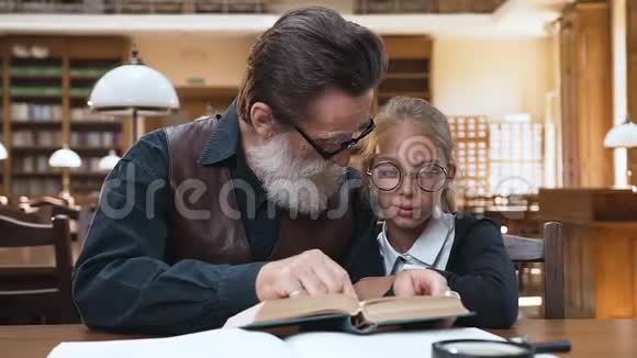 善良的老爷爷留着灰胡子坐在图书馆里和他可爱的小孙女一起看书视频的预览图