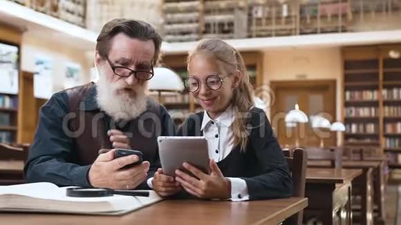 在图书馆里戴着眼镜的尊敬的大胡子老人和他微笑的十几岁的孙女坐在一起视频的预览图