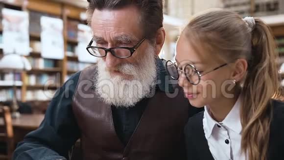 在图书馆里老爷爷和他微笑着的十几岁的孙女一起读有趣的书视频的预览图