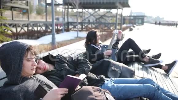 年轻的朋友坐在长凳上不寒而栗两个人在看书一个人在调吉他他的女朋友在看他视频的预览图