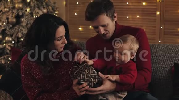 爸爸妈妈和小儿子坐在沙发上在舒适的家背景下在圣诞树附近拿着打开的礼物概念视频的预览图