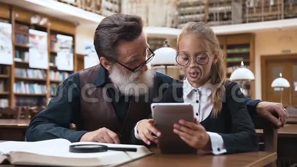 可爱的小女孩戴眼镜阅读故事从电子书到她聪明的大胡子爷爷在图书馆视频的预览图