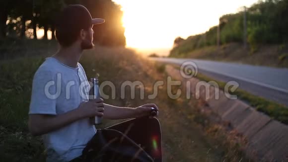 日落时分年轻人坐在草地上喝着钢制热瓶的水靠近沥青路视频的预览图