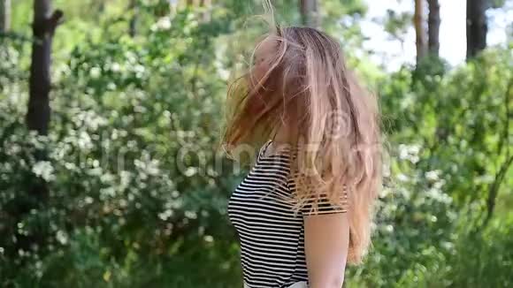 年轻的棕色头发的女人穿着条纹t恤站在阳光下迎着绿色的树叶风吹起她的头发漂亮姑娘视频的预览图
