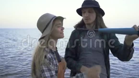 年轻的高加索游客夫妇穿着衬衫和帽子看着望远镜在大海或湖泊的背景下聊天视频的预览图
