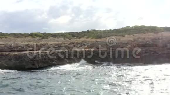 天晴看海浪冲击岩石悬崖海岸在阳光明媚的一天男人坐在岩石上马洛卡岛阳光明媚视频的预览图