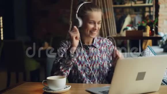快乐的女士享受音乐耳机唱歌跳舞使用笔记本电脑咖啡馆视频的预览图