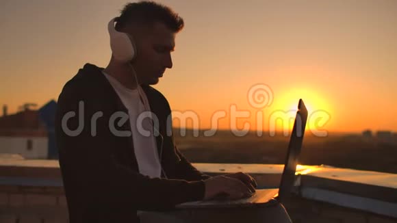 日落时分一个戴着耳机听音乐在建筑物屋顶上工作的人的后视镜视频的预览图