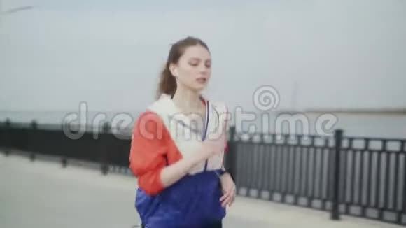 运动的女人沿着河堤奔跑带耳机的运动运动服装中的女孩运动健身模特视频的预览图