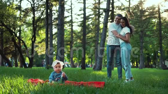 可爱的小女孩坐在绿草地上慈爱的父母从背后看着她视频的预览图