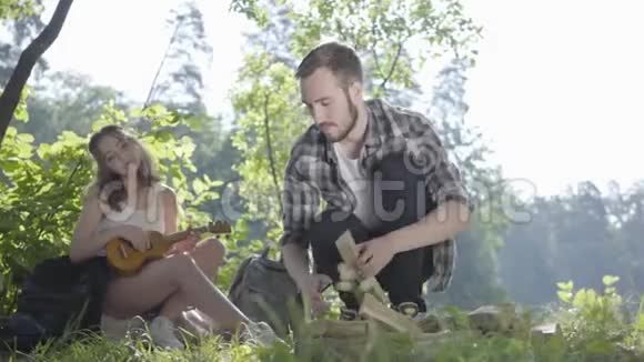 漂亮的年轻女人在背景上玩四弦琴而她的男朋友在森林的前景上生火视频的预览图