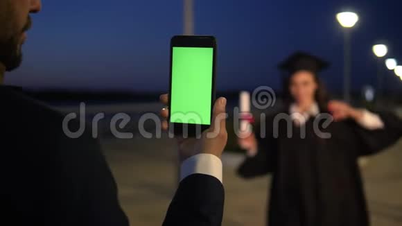 男人拿着绿色屏幕的手机就像在拍一张站在里面的女研究生的照片视频的预览图