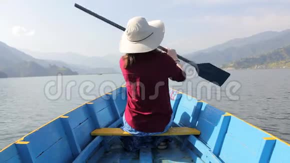 穿着红色衬衫戴着帽子的女孩坐在一艘蓝色的木船上手里拿着桨从后面看着湖靠在后面视频的预览图
