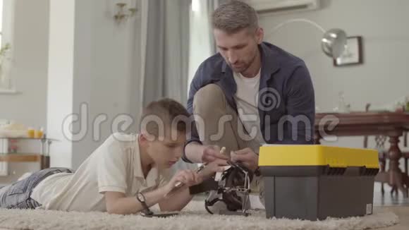 父亲和儿子在家里的地板上铺着毛茸茸的地毯固定着破碎的虚拟现实头盔特写孩子视频的预览图