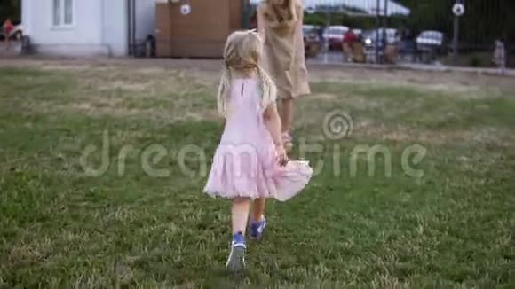 一个漂亮的金发女孩穿着粉红色的连衣裙在草坪上追赶她的母亲一位成年母亲在公园里和她玩耍视频的预览图