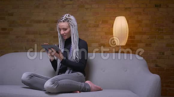 一位年轻迷人的白种人时髦女性在室内沙发上浏览平板短信的特写镜头视频的预览图