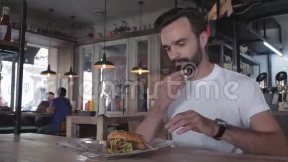 有胡子的时髦小伙子咬着一个汉堡坐在咖啡馆的桌子旁吃汉堡的人4k4k录像60英尺视频的预览图