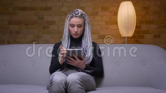 年轻迷人的白种人潮女用平板电脑特写镜头开心地坐在沙发上笑着视频的预览图