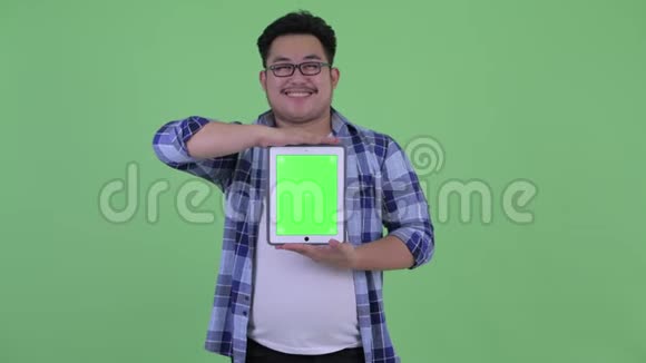 年轻快乐体重超重的亚洲潮人一边思考一边展示数码平板电脑视频的预览图