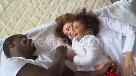 年轻幸福的多民族家庭的母亲非洲父亲和小女儿穿着白色衣服躺在床上视频的预览图