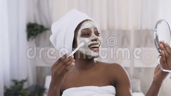 快乐的非裔美国女人头上戴着白色毛巾脸上戴着酸奶面具在镜子里看着健康面部表情视频的预览图