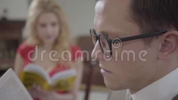 年轻的怪人戴着眼镜看着书在前景中睡着了而金发女郎则在看杂志视频的预览图
