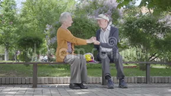 可爱的老妇人拿着一束黄色的花坐在长凳上和一位老人牵着手视频的预览图