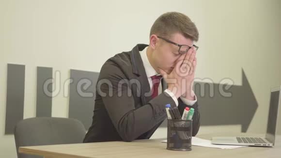 肖像年轻紧张的忙碌的男人穿着正式的衣服戴着眼镜坐在办公室前的笔记本电脑前摩擦他的电脑视频的预览图