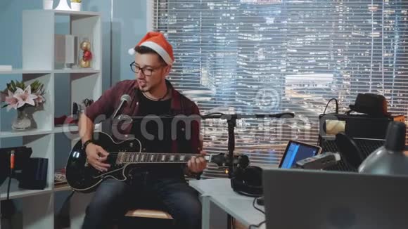 家庭录音室戴着圣诞帽的年轻歌手用麦克风和吉他唱圣诞歌曲视频的预览图