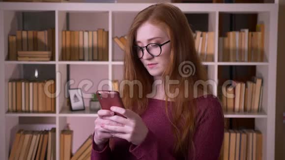 戴着眼镜的年轻红发女孩的特写肖像戴着手机的白种人女学生开心地笑着视频的预览图