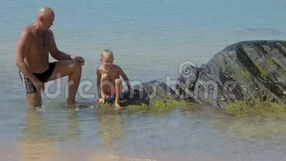 小男孩坐在大海上快乐父亲附近的大石头上视频的预览图