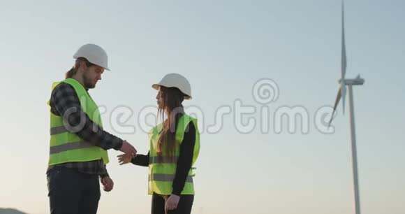 工程师们戴着头盔的男人和女人在风车涡轮背景上握手视频的预览图