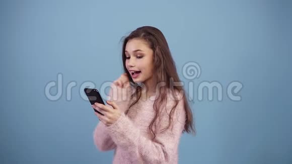 穿着粉色毛衣的快乐少女使用她的智能手机发短信在工作室里以蓝色背景隔绝视频的预览图
