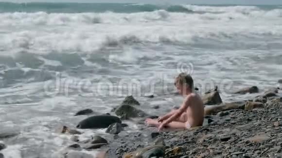 男孩坐在海浪中在石滩上的暴风雨中玩耍后景视频的预览图